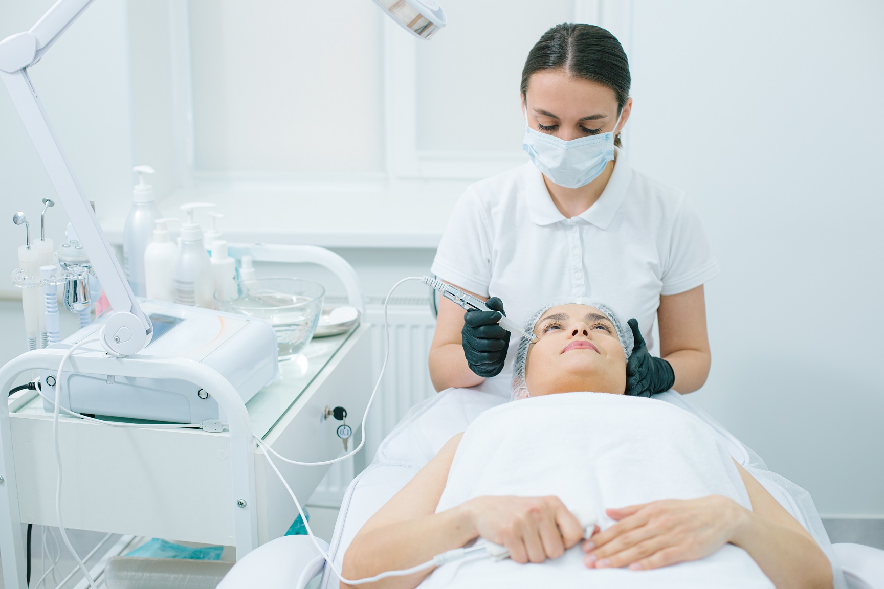 Beautician in medical mask touching the face of her female client with metal prongs during the ultrasonic cleansing